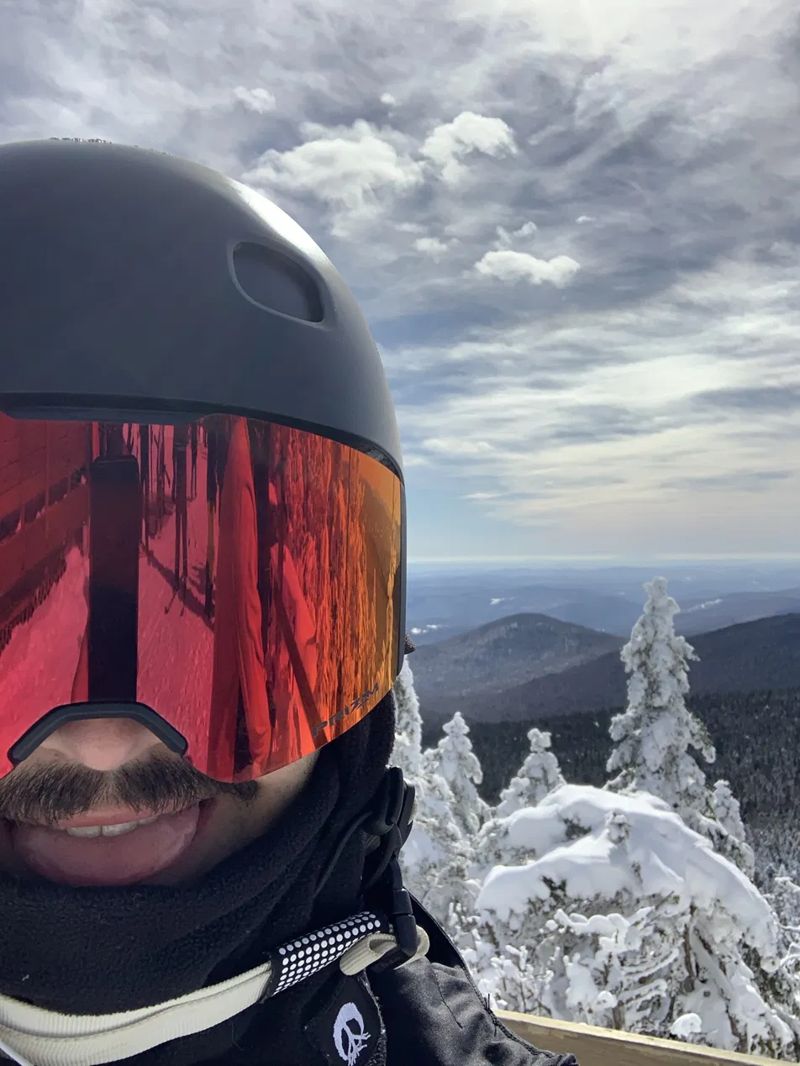 Ski selfie with mirrored goggles above snow-covered trees and mountains